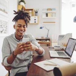 年轻的黑人男子穿着衬衫，坐在电脑前对着电话笑